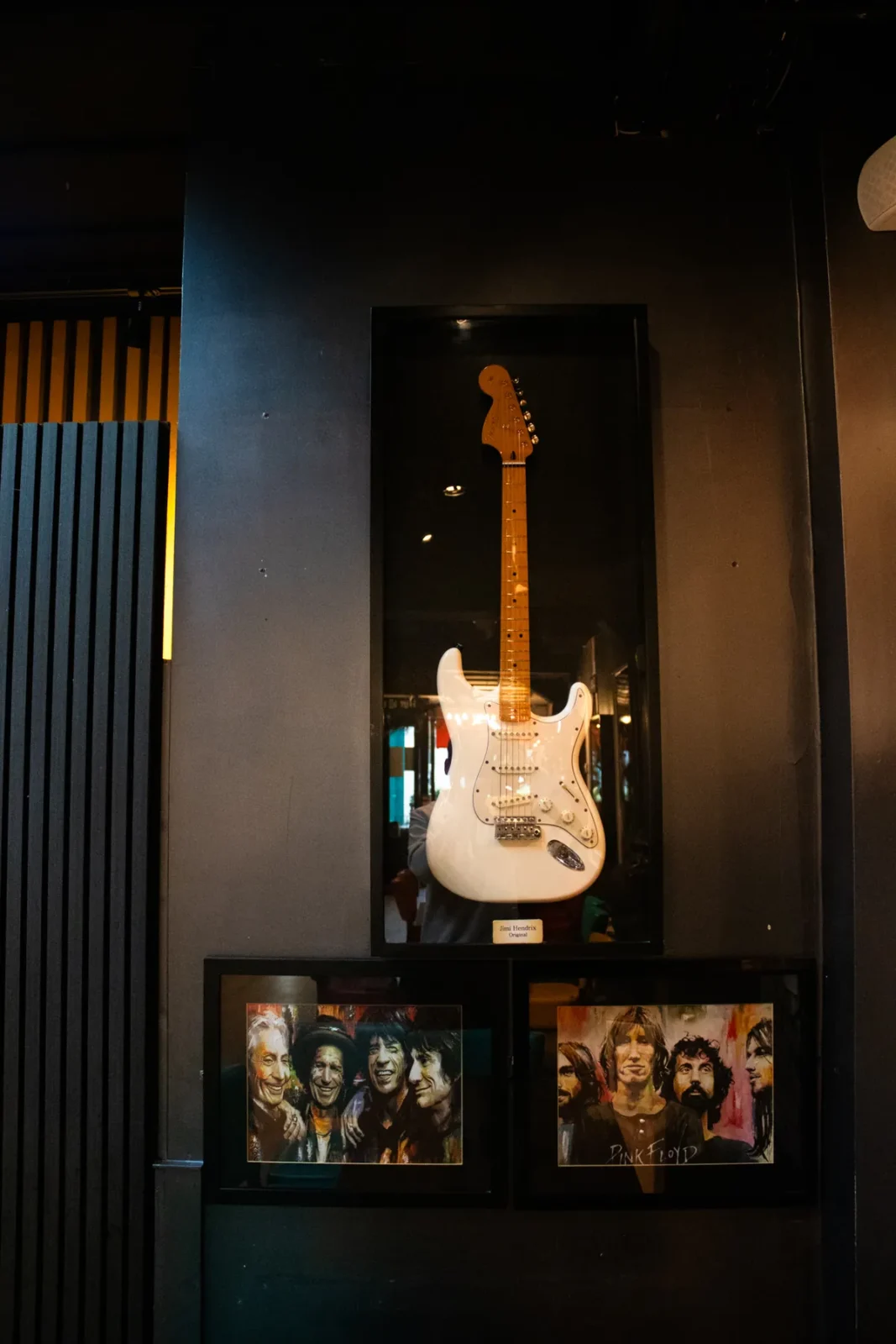 A white electric guitar in its own case, hung above two framed band photos.