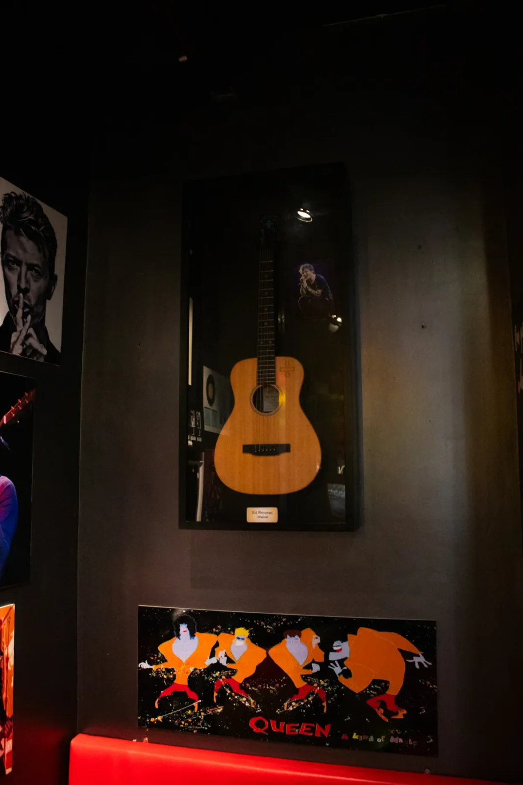 An acoustic guitar in a glass case, with a Queen print underneath.
